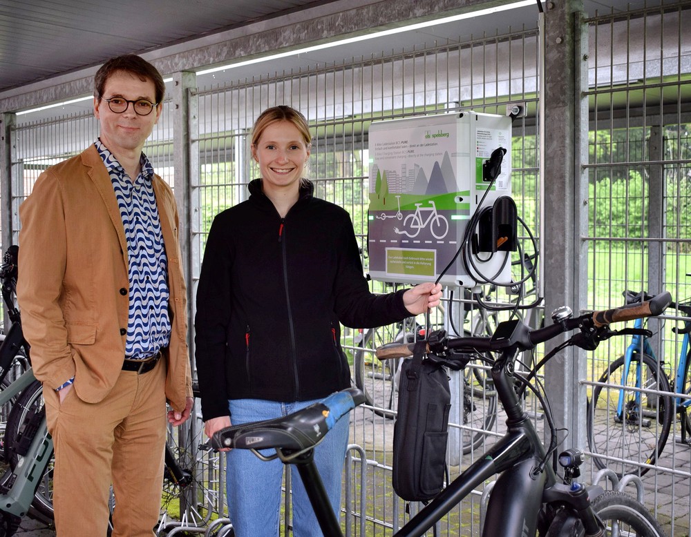 Das Bild zeigt eine Frau und einen Mann, die an einer Fahrradladestation stehen.