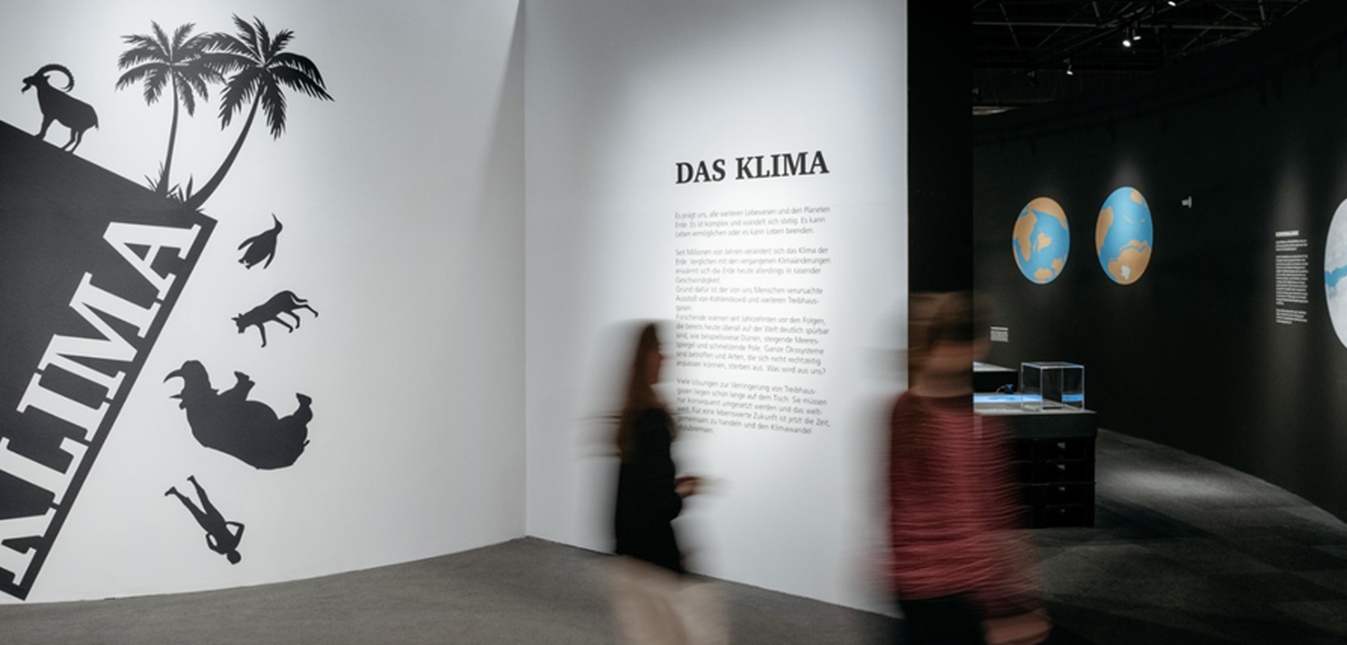 Ein Raum im Muesum: Im Hintergrund hängen Bilder, im Vordergrund gehen zwei Menschen an einer beschrifteten Wand vorbei.