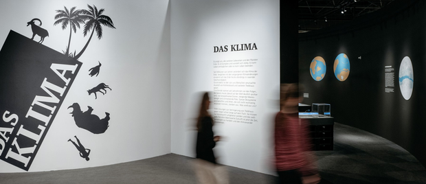 Das Bild zeigt einen Raum im Museum. Im Hintergrund hängen Bilder, im Vordergrund gehen zwei Menschen an einer beschrifteten Wand vorbei.
